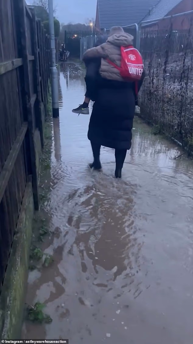 Parents carry their children to school through muddy water ’caused by monstrous 60ft tall warehouse’ – as council promises to dig a ditch ‘as soon as weather permits’